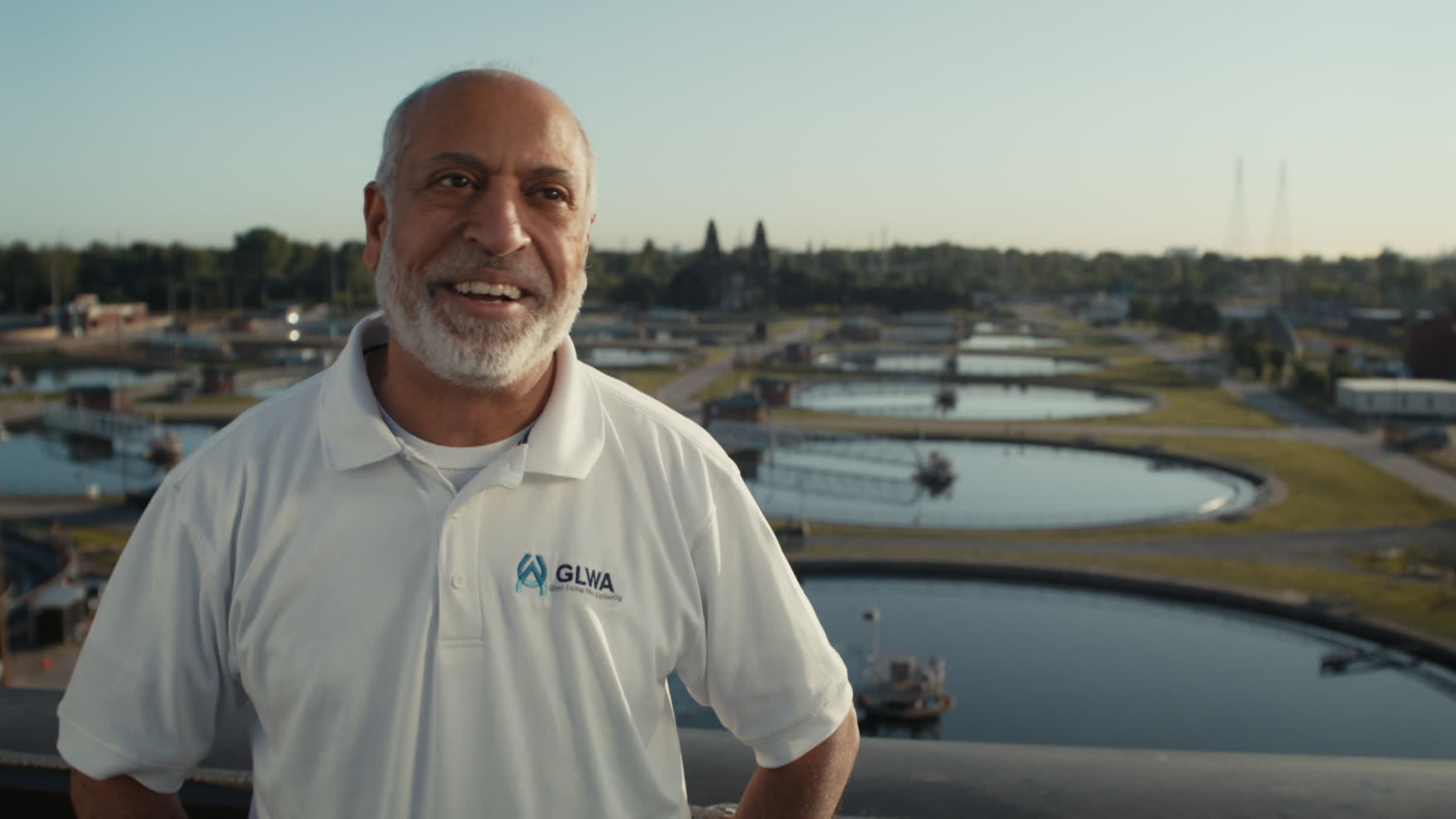 A man, smiling, stands in front of a water treatment facility. 