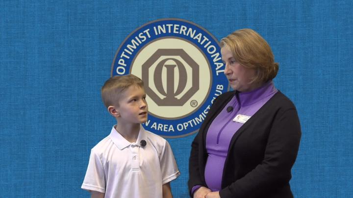 A boy, left, speaks to a representative from the Clarkston Area Optimist Club with the club's logo behind them on a blue background. 