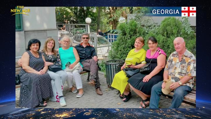 Seven people sitting on a bench in the country Georgia, flag in upper right corner, with four people to the left and three to the right. 