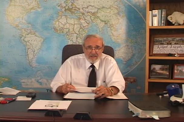 A man sitting center with a map behind him and a bookcase to the right. 