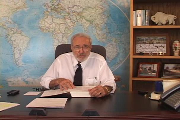 A man sitting center with a map behind him and a bookcase to the right. 