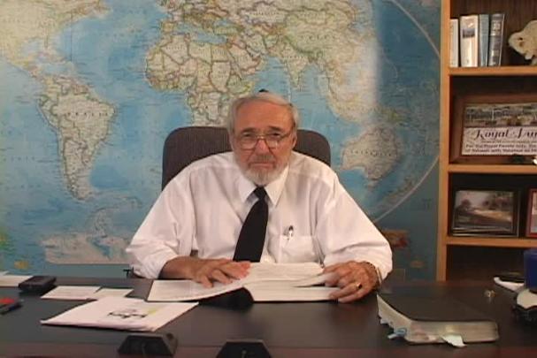 A man sitting center with a map behind him and a bookcase to the right. 