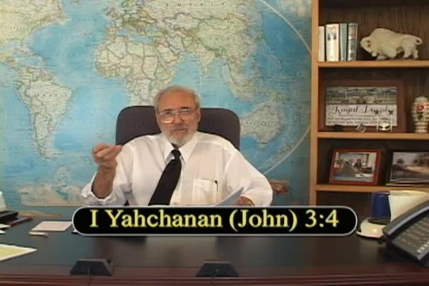 A man gestures with his right hand while sitting center with a map behind him and a bookcase to the right. 