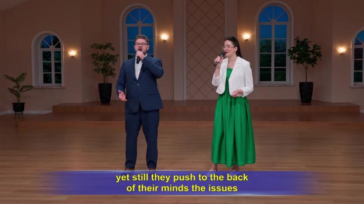 A man, left, and woman, right, singing in a room with four windows behind them. 