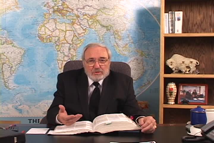 A man sitting center with a map behind him and a bookcase to the right. 