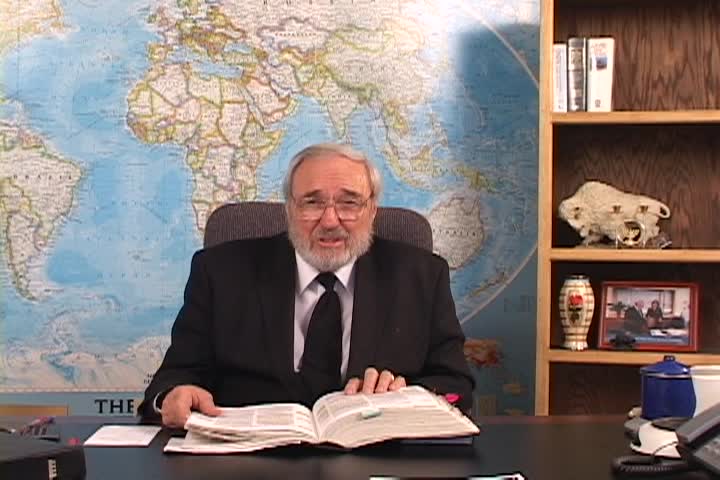 A man sitting center with a map behind him and a bookcase to the right. 