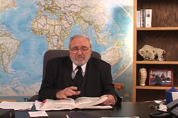 A man sitting center with a map behind him and a bookcase to the right. 
