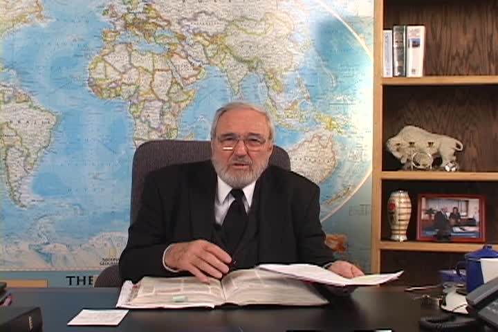 A man sitting center with a map behind him and a bookcase to the right. 