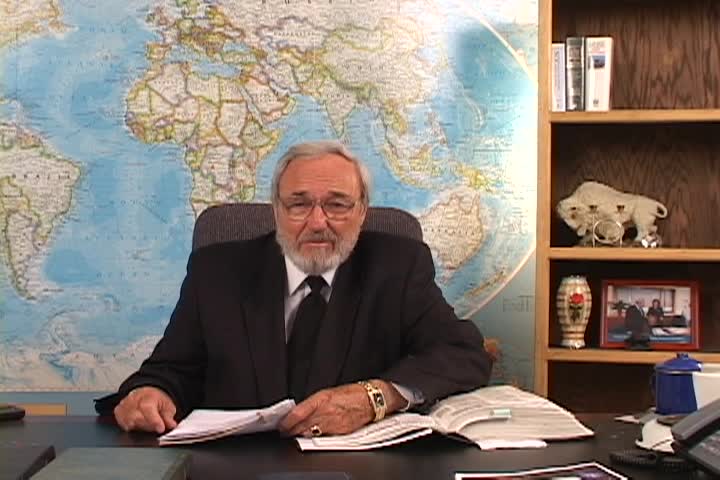 A man sitting center with a map behind him and a bookcase to the right. 
