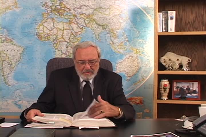 A man sitting center with a map behind him and a bookcase to the right. 