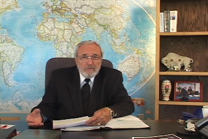 A man sitting center with a map behind him and a bookcase to the right. 