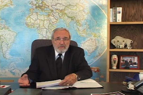 A man sitting center with a map behind him and a bookcase to the right. 