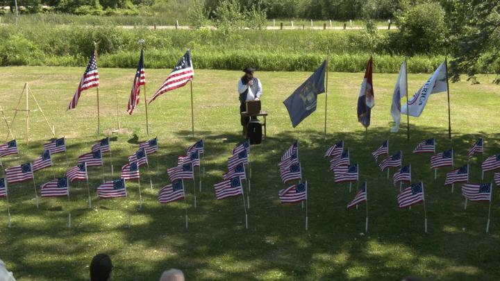 A man stands at a podium, center frame, surrounded by flags. 